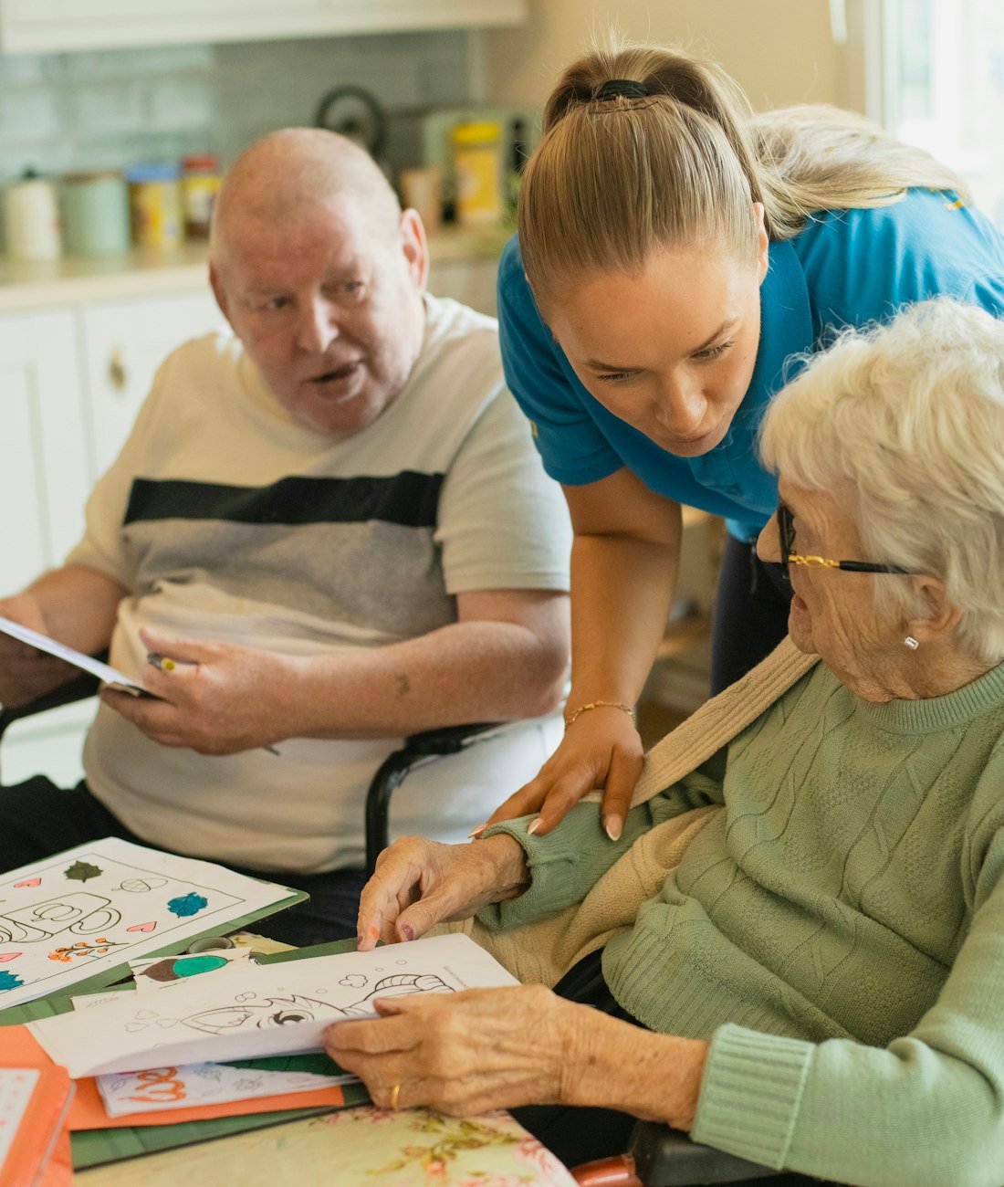 A caregiver assisting an elderly couple with an activity at their dining table