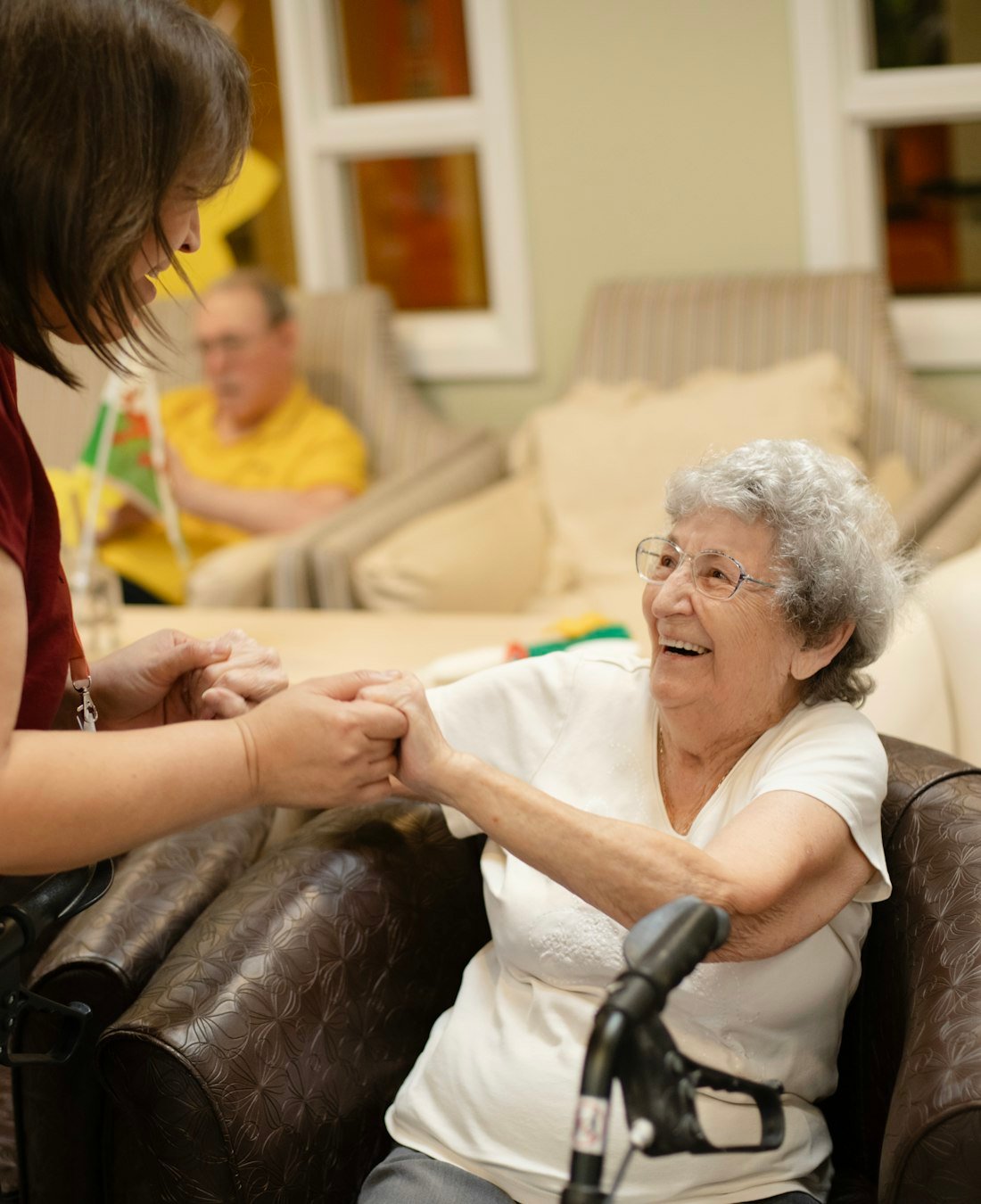 A caregiver holding the hands of a smiling elderly woman in her living room